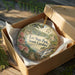 A decorative ornament in a box, featuring a scenic landscape with a waterfall and a bridge.