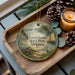 A decorative ornament featuring a scenic image of LESS THAN 500 characters, including a house, boat, and pine cones, placed on a wooden tray alongside a lit candle.