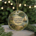 A round ornament with a painting of a sailboat and a cabin, surrounded by greenery, hangs in front of a Christmas tree with lights.