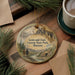 A decorative ornament featuring a scenic landscape with mountains, pine trees, and a river, hanging on a wooden surface.
