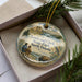 A decorative ornament in a box, featuring a scenic landscape with people, animals, and natural elements.