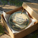 A circular ornament with a landscape scene and text, placed in a cardboard box with a gold string.
