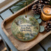 A decorative ornament featuring a scenic landscape of Lincolumbus Woods State Park, Rhode Island, with pine cones, a candle, and greenery surrounding it.