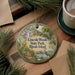 A decorative ornament featuring a scenic landscape of Lincolns Woods State Park, Rhode Island, with a bird and a waterfall. The ornament is placed on a wooden surface, surrounded by pine branches and a cup of coffee.