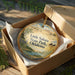 A decorative ornament in a box, featuring a scenic landscape with a body of water and mountains in the background.