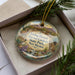 A decorative ornament with a desert landscape and wildlife scene, featuring a bird, a cat, and cacti, is displayed in a box with pine branches.