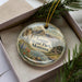 A decorative ornament in a box, featuring a scenic landscape with mountains, water, and pine trees.