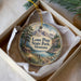 A decorative ornament in a box, featuring a scenic landscape with mountains, pine trees, and a cabin.