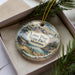 A decorative ornament in a box, featuring a scenic landscape with mountains, pine trees, and a cabin.