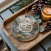 A wooden tray with a decorative ornament featuring a scenic landscape of mountains, pine trees, and a cabin, along with pine cones and a lit candle.