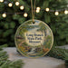 A decorative ornament featuring a scenic landscape with a deer, a waterfall, and a cabin, hanging in front of a Christmas tree with lights.