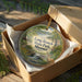 A decorative ornament in a box, featuring a scenic landscape with a deer and a waterfall.