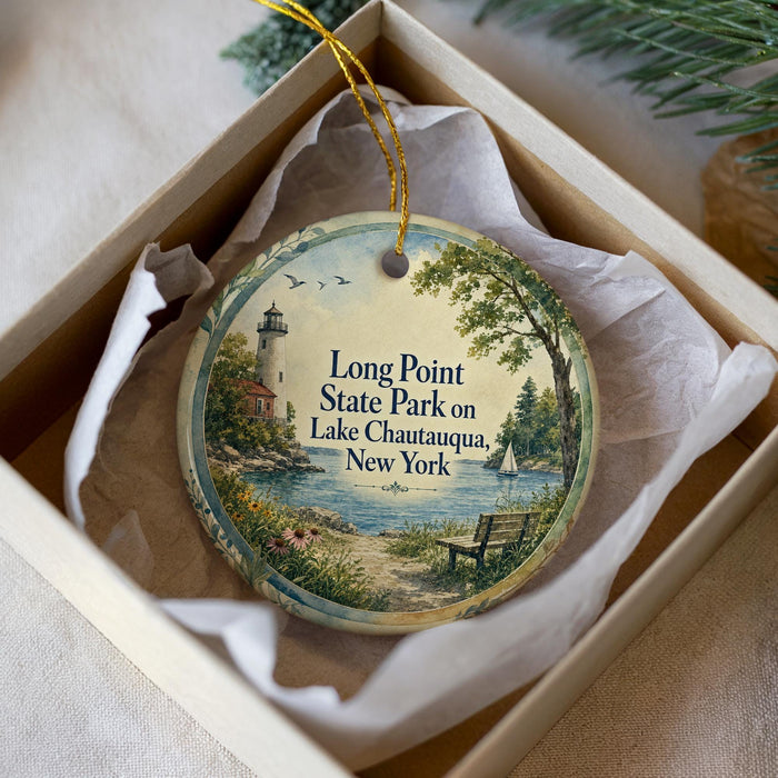 A decorative ornament in a box, featuring a lighthouse and a scenic landscape with a lake and a bench.