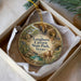 A circular ornament with a painting of a scenic landscape, including a cave, trees, and a cow, is displayed in a box.
