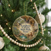 A circular ornament hanging on a Christmas tree, featuring an illustration of a scenic landscape with a large rock formation and a cow in the foreground.