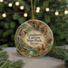 A circular ornament with a painting of a scenic landscape, featuring a cow, trees, and a pond, hanging in front of a Christmas tree with lights.