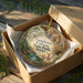 A circular ornament with a landscape scene inside, placed in a cardboard box with tissue paper.