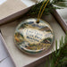 A decorative ornament in a box, featuring a scenic landscape with mountains, trees, and a stream.