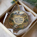 A decorative ornament in a box, featuring a scenic landscape with mountains, trees, and a stream.