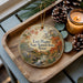 A wooden tray with a decorative ornament featuring a scenic landscape of the Lost Maples State Natural Area in Texas, accompanied by pine cones and a lit candle.