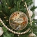 A circular ornament hanging on a Christmas tree, featuring a scenic landscape with a river, mountains, and fall foliage.