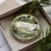 A decorative ornament in a box, featuring a scenic landscape with a river, mountains, and greenery.