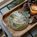 A wooden tray with a decorative ornament featuring a scenic landscape of the Louisville State Recreation Area in Nebraska, accompanied by pine cones and a lit candle.