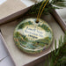 A decorative ornament in a box, featuring a scenic landscape with a river, bridge, and trees.