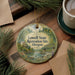 A round ornament with a scenic illustration of a river and bridge, surrounded by pine branches, sits on a wooden surface.