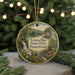 A circular ornament featuring a scenic landscape with a waterfall, pine trees, and a bridge, hanging in front of a blurred background with Christmas lights.