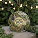 A round ornament featuring a scenic landscape with a waterfall, trees, and a bridge, hanging in front of a Christmas tree with lights.