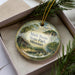 A decorative ornament in a box, featuring a scenic landscape with a cabin and a river.