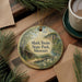 A round ornament with a painting of a scenic landscape hangs on a wooden surface, surrounded by pine branches and a cup of coffee.