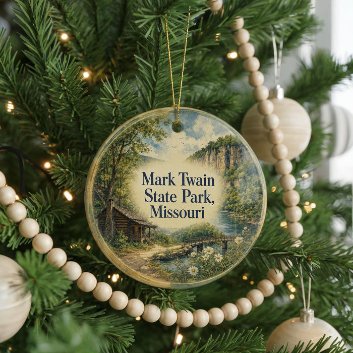 A round ornament hanging on a Christmas tree, featuring a scenic landscape with a cabin and a river.