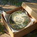 A circular ornament in a box, featuring a scenic landscape with a cabin and trees, with the text "Mark Twain State Park, Missouri" printed on it.