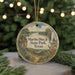 A round ornament with a painting of a scenic landscape, featuring a waterfall, trees, and a deer, hangs in front of a Christmas tree with lights.