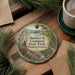A round ornament with a scenic image of a park, featuring a river, mountains, and pine trees, hangs on a wooden surface.