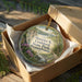 A round ornament in a brown box, featuring a scenic landscape with a river and mountains, is displayed on a wooden surface.