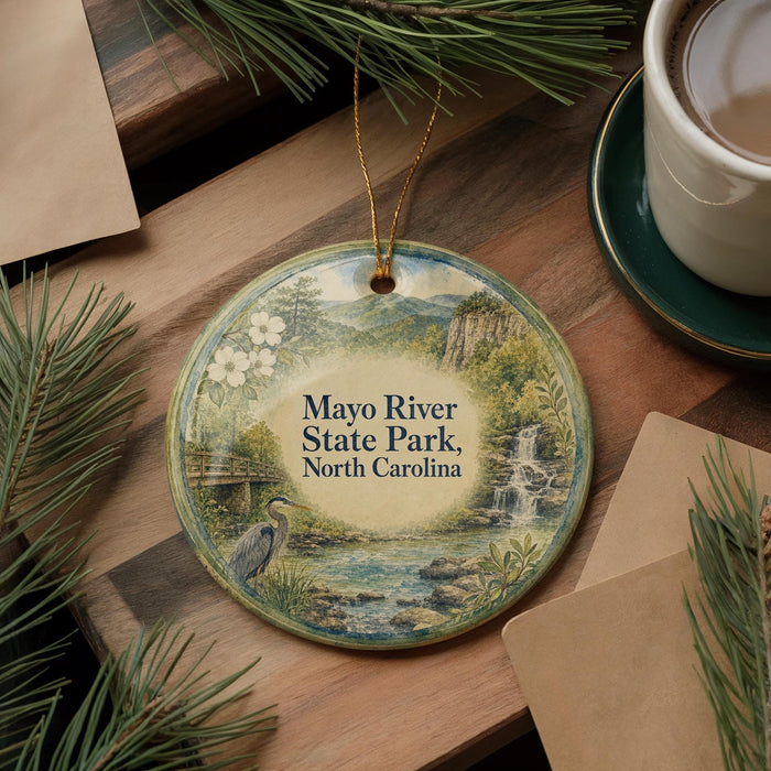 A round ornament with a scenic image of the Mayo River State Park in North Carolina, featuring a bird, a waterfall, and a bridge. The ornament is hanging from a string and is placed on a wooden surface, surrounded by pine branches and a cup of coffee.