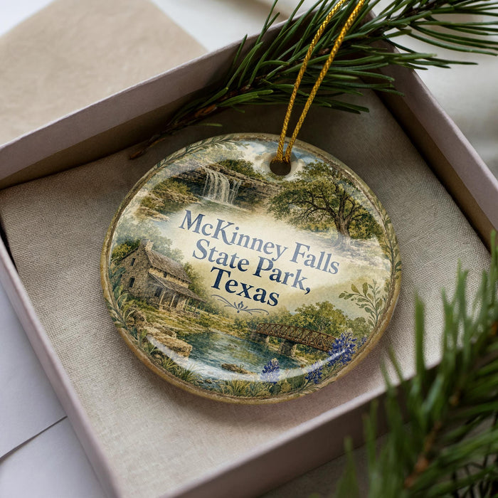 A decorative ornament with a scenic image of McKinney Falls State Park, Texas, displayed in a box with pine branches.