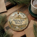 A round ornament with a scenic mountain landscape and text, hanging on a wooden surface next to a cup of coffee and pine branches.
