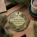 A decorative ornament with a scenic forest landscape, featuring a deer, a bird, and a canoe, hangs on a wooden surface alongside a cup of coffee and some pine branches.