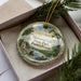 A decorative ornament in a box, featuring a scenic landscape with a lake, mountains, and a castle.