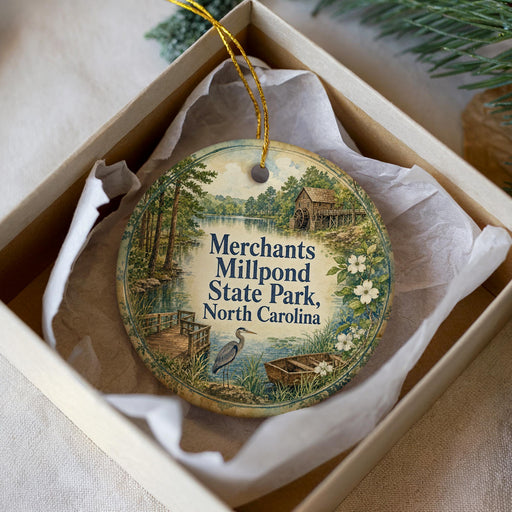 A decorative ornament in a box, featuring a scenic landscape with a lake, a bridge, and a bird, along with text that reads "Merchants Millpond State Park, North Carolina".
