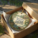 A decorative ornament in a wooden box, featuring a scenic landscape with a bird and a house, with the text "Merchants Millpond State Park, North Carolina" printed on it.