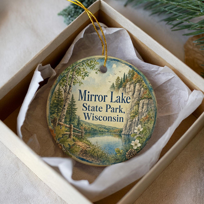 A round ornament with a scenic landscape of Mirror Lake State Park, Wisconsin, displayed in a box with a white tissue paper lining.