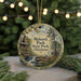 A circular ornament with a painting of a cabin and a waterfall, surrounded by greenery, hangs in front of a blurred background with twinkling lights.