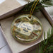 A decorative ornament with a scenic image of Mohican State Park, Ohio, displayed in a box with pine branches.