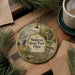 A circular ornament with a scenic illustration of Mohican State Park, Ohio, featuring a waterfall, cabin, and trail signs, is displayed on a wooden surface alongside a cup of coffee and some pine branches.