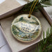A decorative ornament in a box, featuring a scenic landscape with a river, trees, and mountains.
